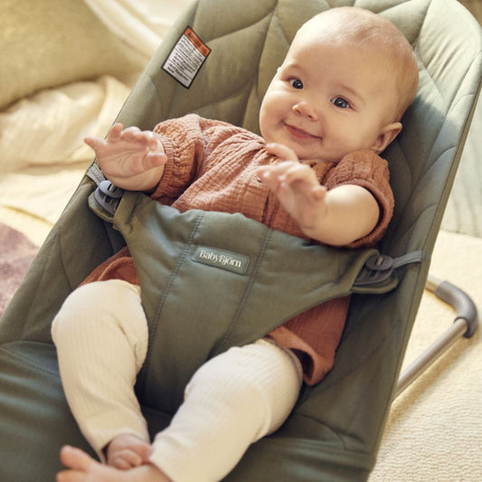 Baby in a green BabyBjorn baby bouncer with a visible brand logo, sitting on a beige surface.