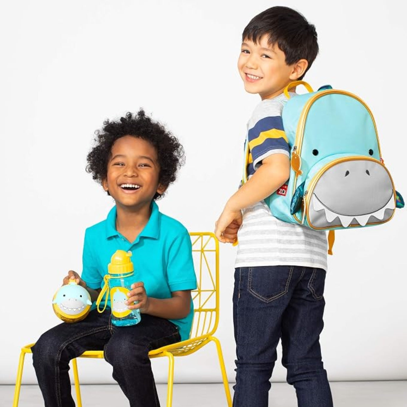 Two children in a bright studio setting. One child stands wearing a light blue shark-shaped Skip Hop backpack with a gray mouth and yellow trim, while the other sits on a yellow chair holding a matching light blue and yellow water bottle and a small shark-themed item.