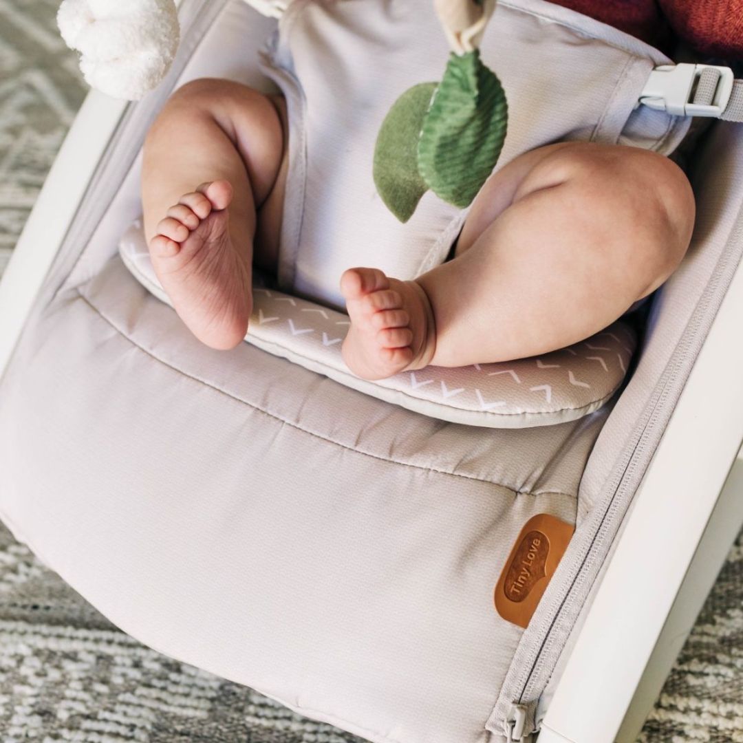 Baby in a white outfit with green leaf toy inside a gray Tiny Love baby bouncer.