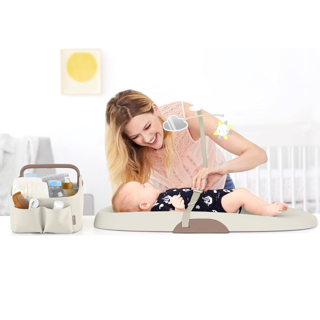 A mother smiling and adjusting a mobile toy above her baby lying on a changing pad, with the filled Skip Hop diaper caddy positioned nearby on the table.