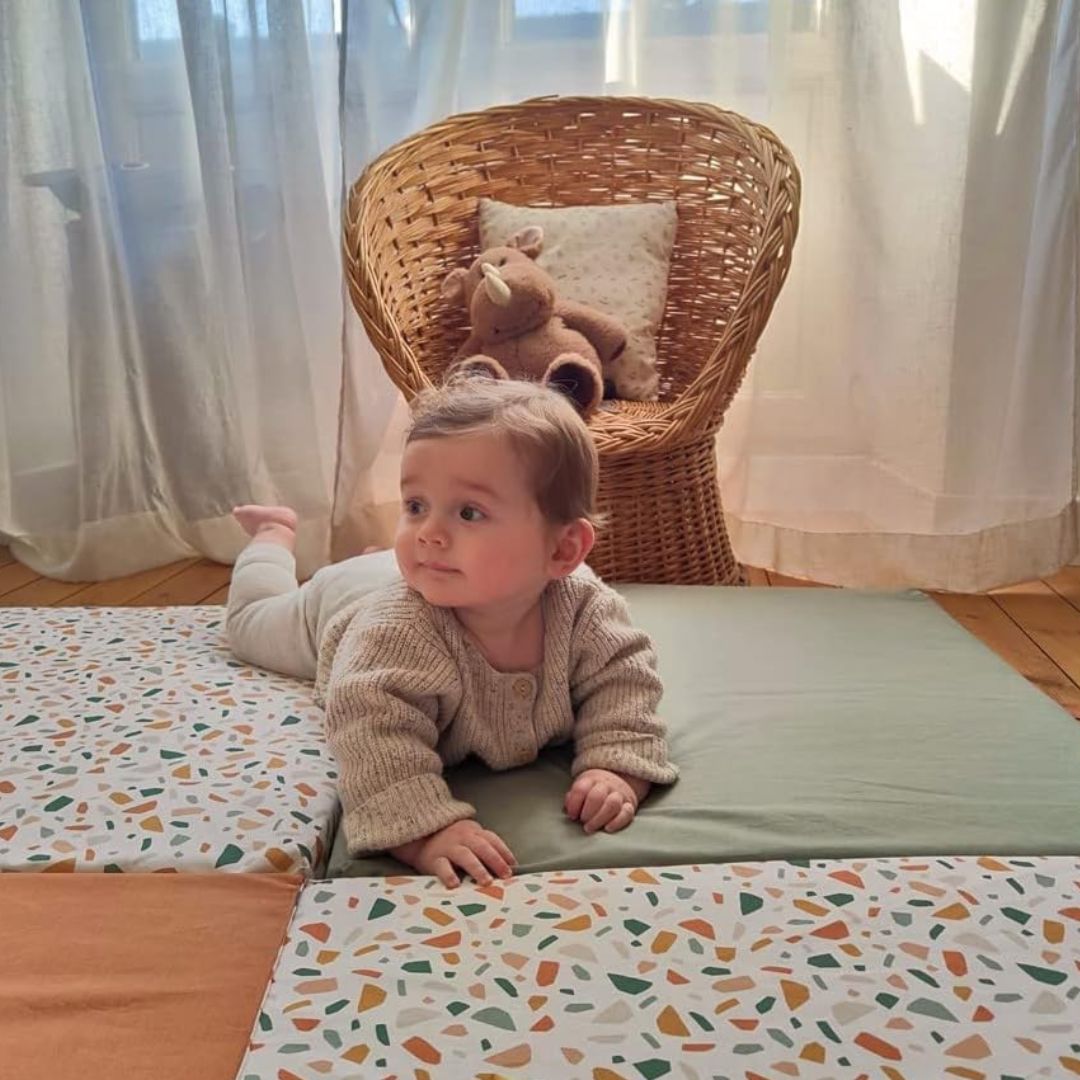 Baby lying on a colorful candide mattress with a wicker chair and teddy bear in the background.
