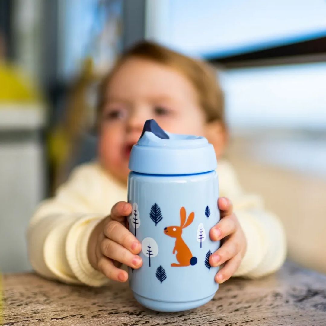 Child holding a blue sippy tommee tippee cup with animal illustrations in a kitchen setting