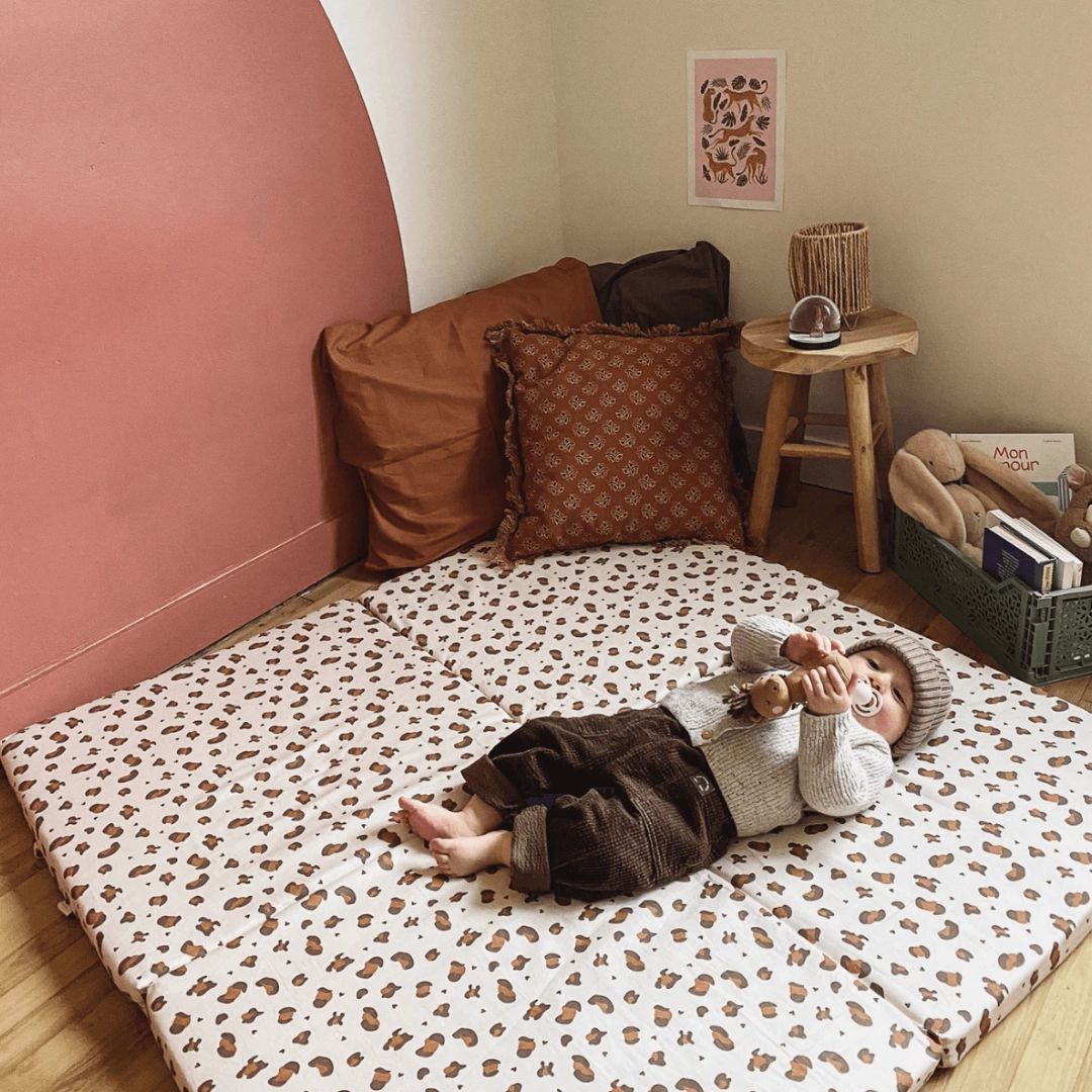 Child lying on a leopard print mat in a cozy room with pillows and a small table.