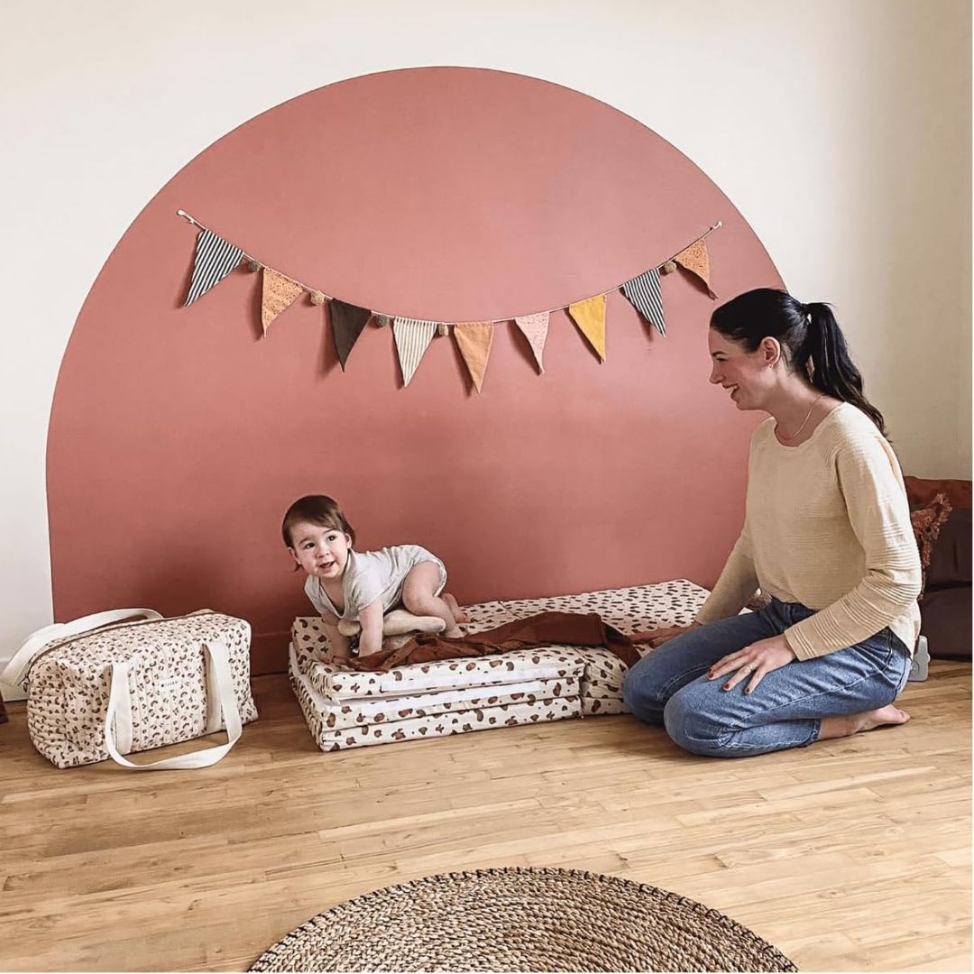 Woman and child playing on a patterned mat with a pink wall and decorative flags in the background.