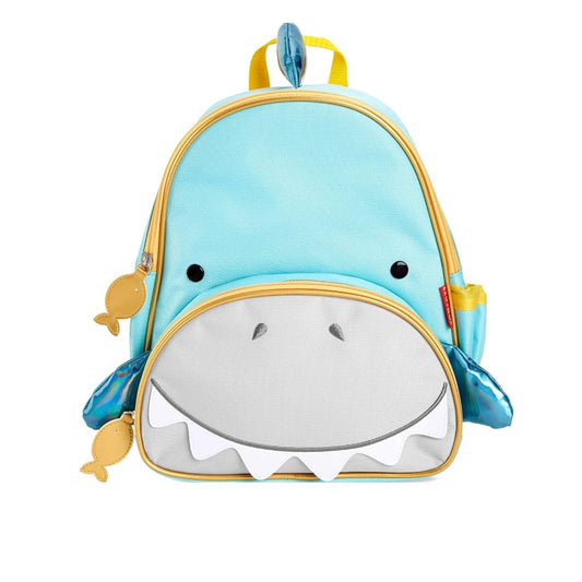 A Skip Hop light blue shark-shaped children's backpack with a white mouth and teeth front design, yellow zipper trim, and small yellow fish charms on the sides, shown on a plain white background.