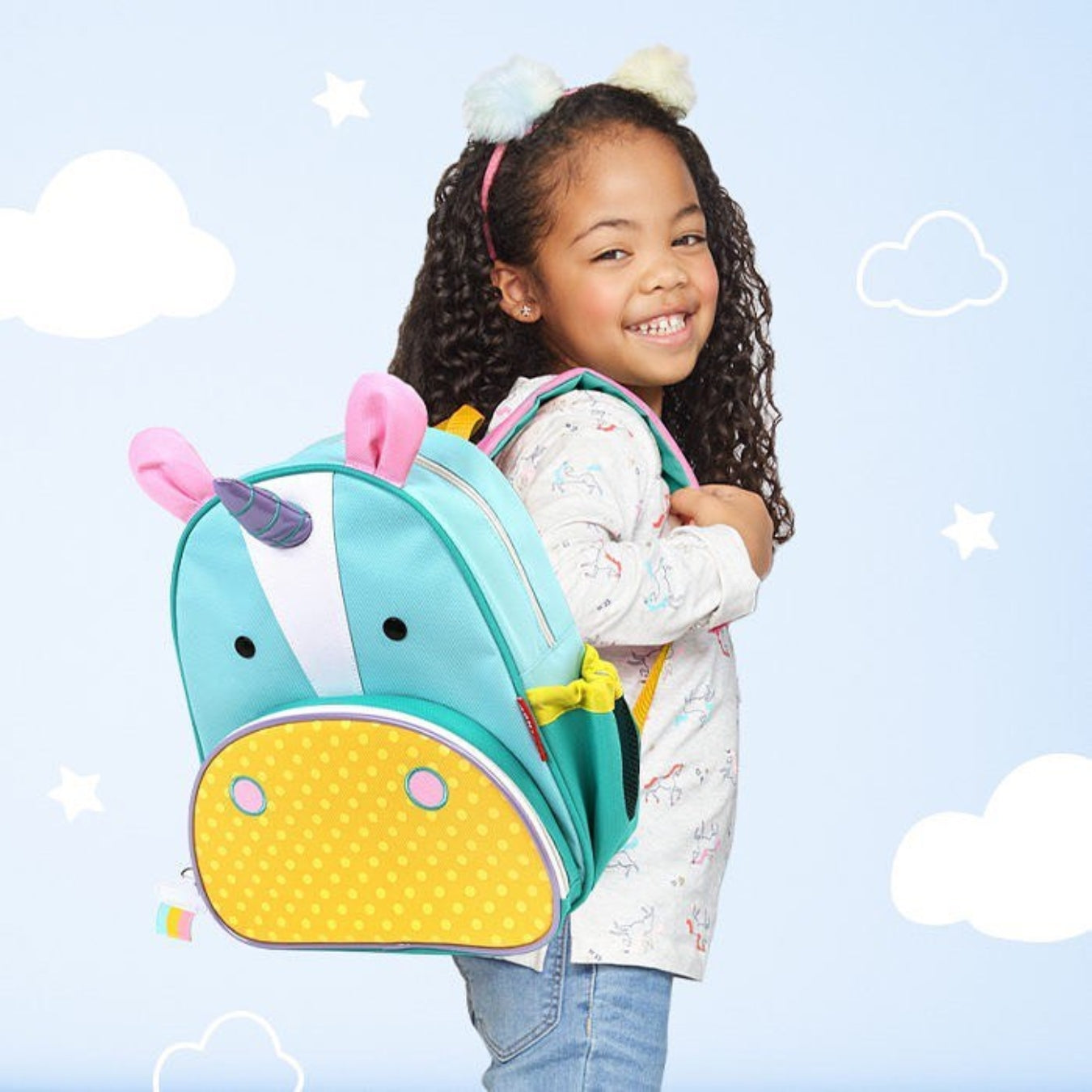 A smiling young girl with curly hair wearing a Skip Hop unicorn-themed backpack. The backpack is light blue with a yellow snout, pink ears, and a purple horn, shown against a light blue background with white clouds and stars.