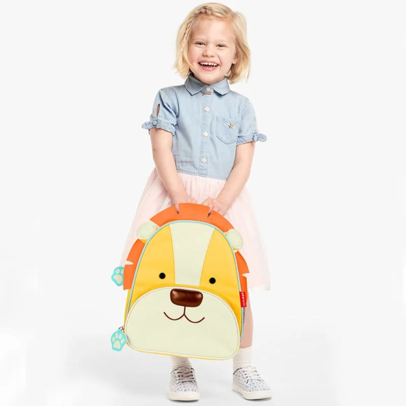 A young girl with blonde hair smiling and holding a colorful lion-face backpack. She is wearing a light blue short-sleeve button-up shirt, a light pink skirt, white socks, and white sneakers, set against a plain white studio background.