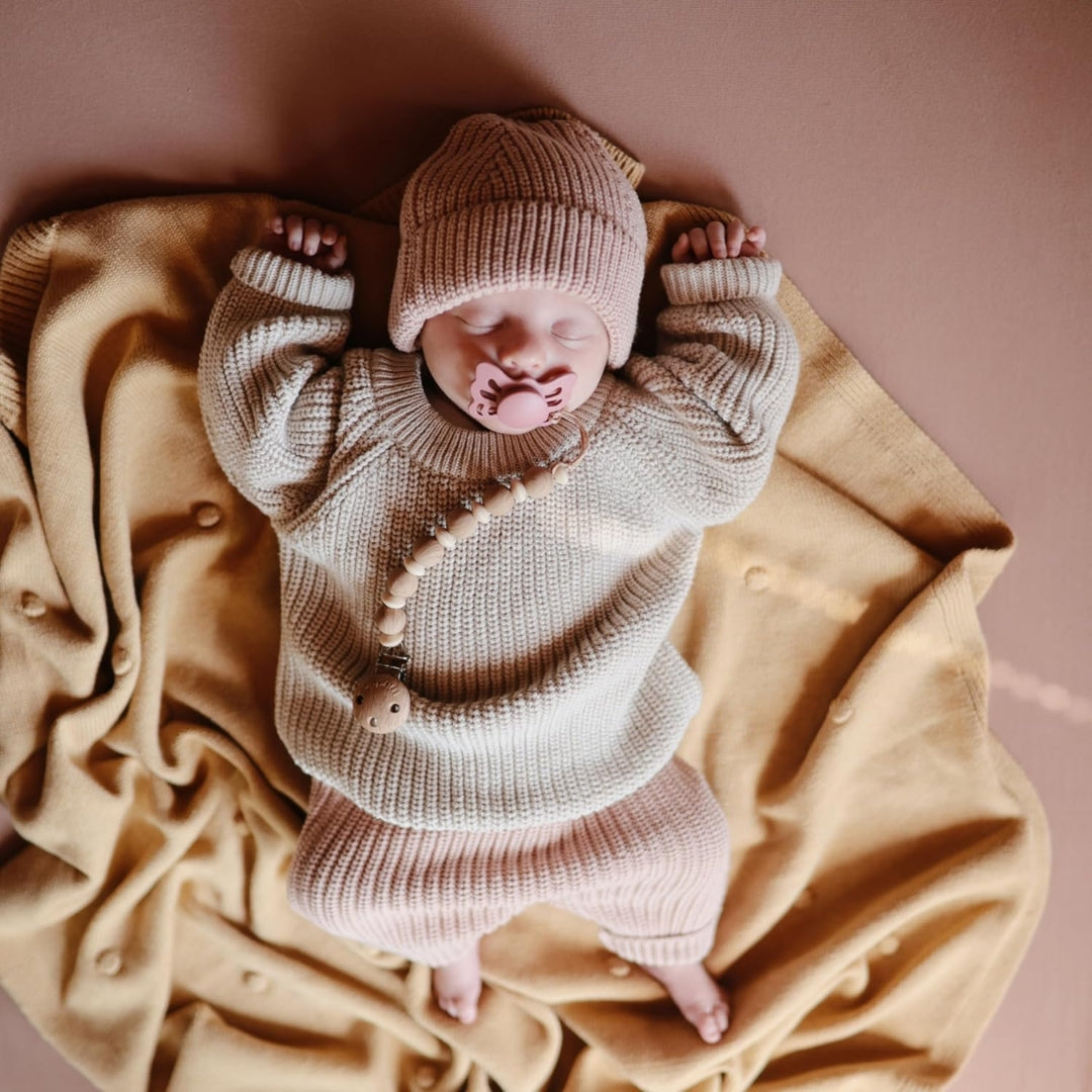 Newborn baby in a knitted outfit and hat lying on a beige blanket with a frigg pacifier