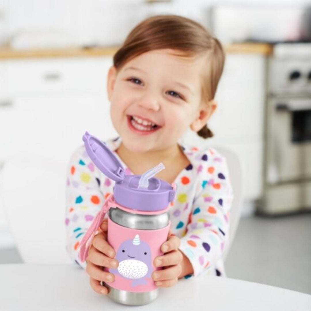 A young child smiling while holding a pink and purple insulated Skip Hop water bottle with a unicorn design, set against a softly blurred kitchen background.