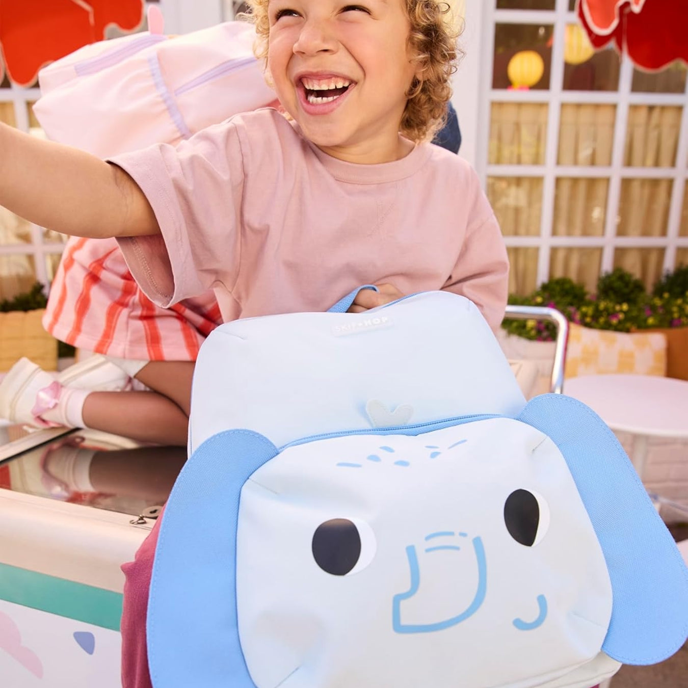 A smiling child in a pink shirt sits behind a large light blue and Light blue Skip Hop character backpack with floppy ears and a cartoon face. The scene appears to be a playful indoor setting with bright decor in the background.