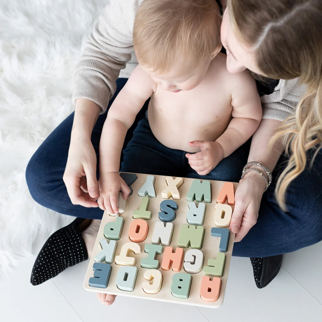 Baby playing with a Pearhead wooden alphabet puzzle held by an adult on a white floor.