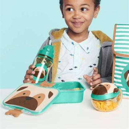 A child wearing a white polo shirt with a blue fish pattern and a gray zip hoodie stands behind a dog-themed lunch set. The set includes a Skip Hop lunchbox with a dog face lid, a matching water bottle, and a striped lunch bag, all in a bright studio setting with a light blue background.