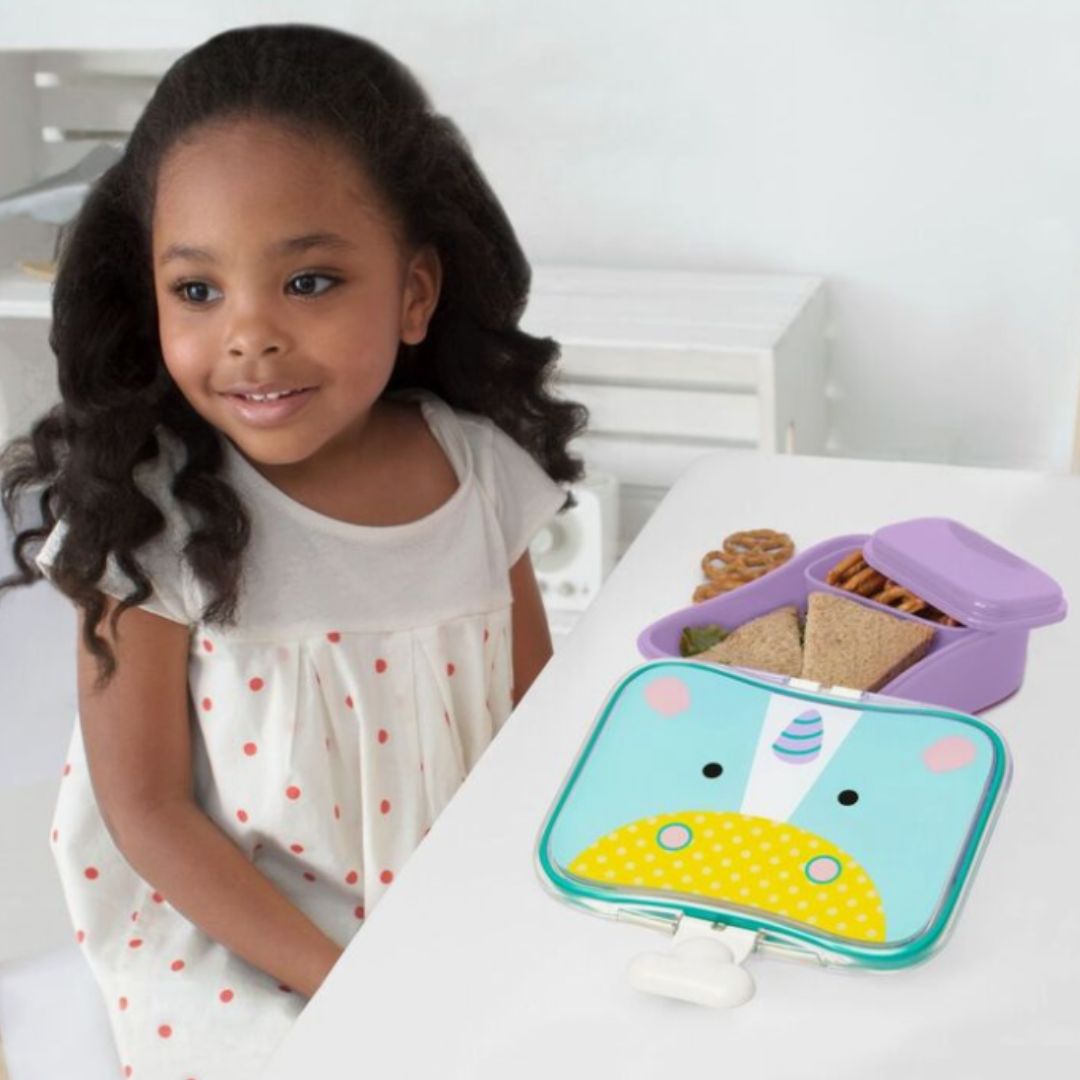 A young girl with dark curly hair wearing a white dress with red polka dots sits at a white table next to a colorful animal-themed Skip Hop lunch box. The lunch box features a light blue and yellow dinosaur face design with a purple lid, containing sandwiches and cookies.