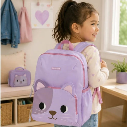 A young girl with a ponytail is shown from behind wearing a light purple cat-face Skip Hop backpack with pink accents in a bright room. The brand name 'Skip Hop' is visible on the front of the backpack.