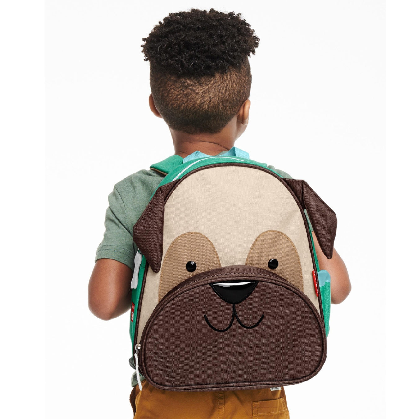 A child viewed from behind wearing a dog-face backpack. The backpack is cream and brown with a black nose and mouth detail, and teal shoulder straps, shown against a plain white background.