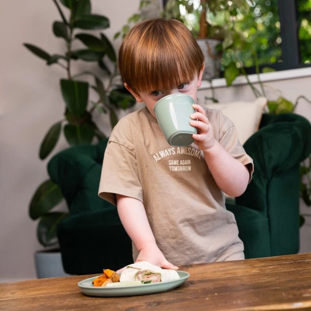 Child drinking from a mug with a plate of food on a table, surrounded by plants.