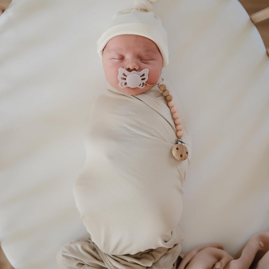 Newborn baby wrapped in a white blanket with a Frigg pacifier and teething ring.