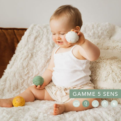 A baby sitting on a fluffy cream blanket holding a white Sophie la Girafe musical ball up to their ear, with a green and yellow ball nearby, alongside a '5 senses range' icon graphic.