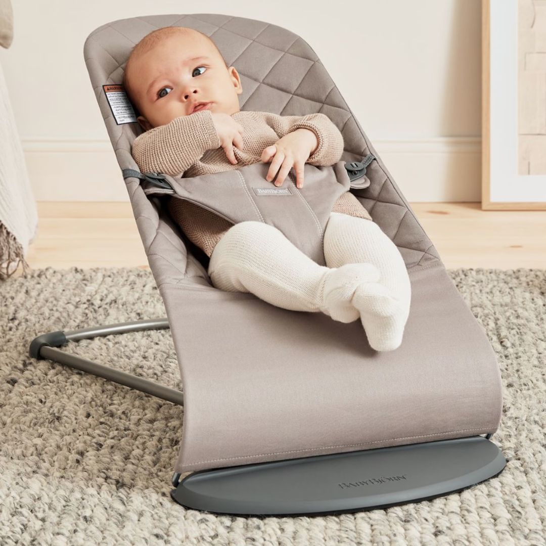 Baby Jorn Baby in a beige baby bouncer on a carpeted floor