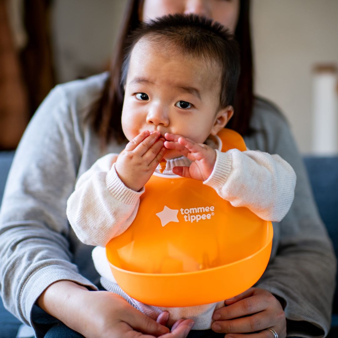 Child wearing an orange bib with 'Tommee Tippee' logo, held by a person.