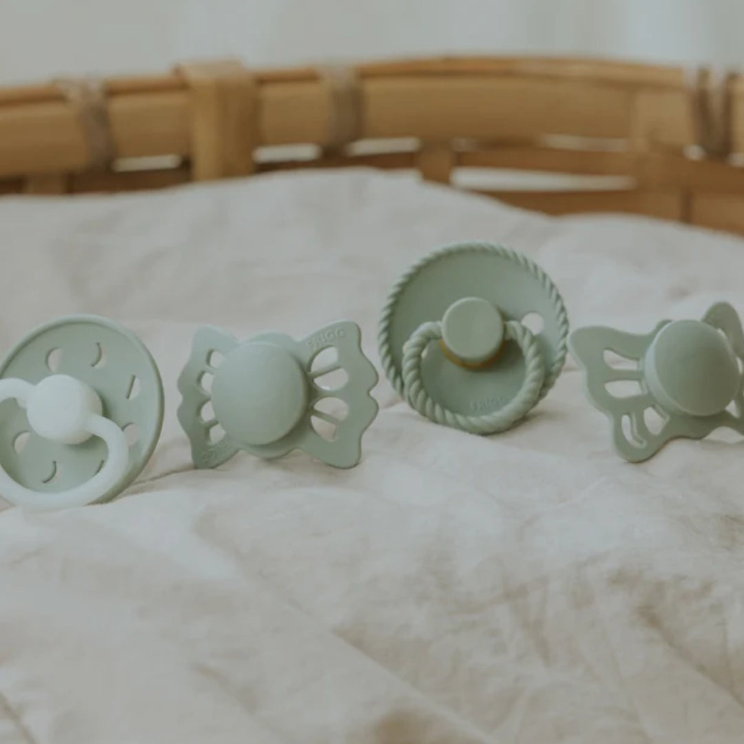 Set of four green baby soothers on a white surface with a wooden basket in the background.