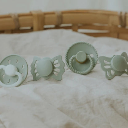 Set of four green baby soothers on a white surface with a wooden basket in the background.