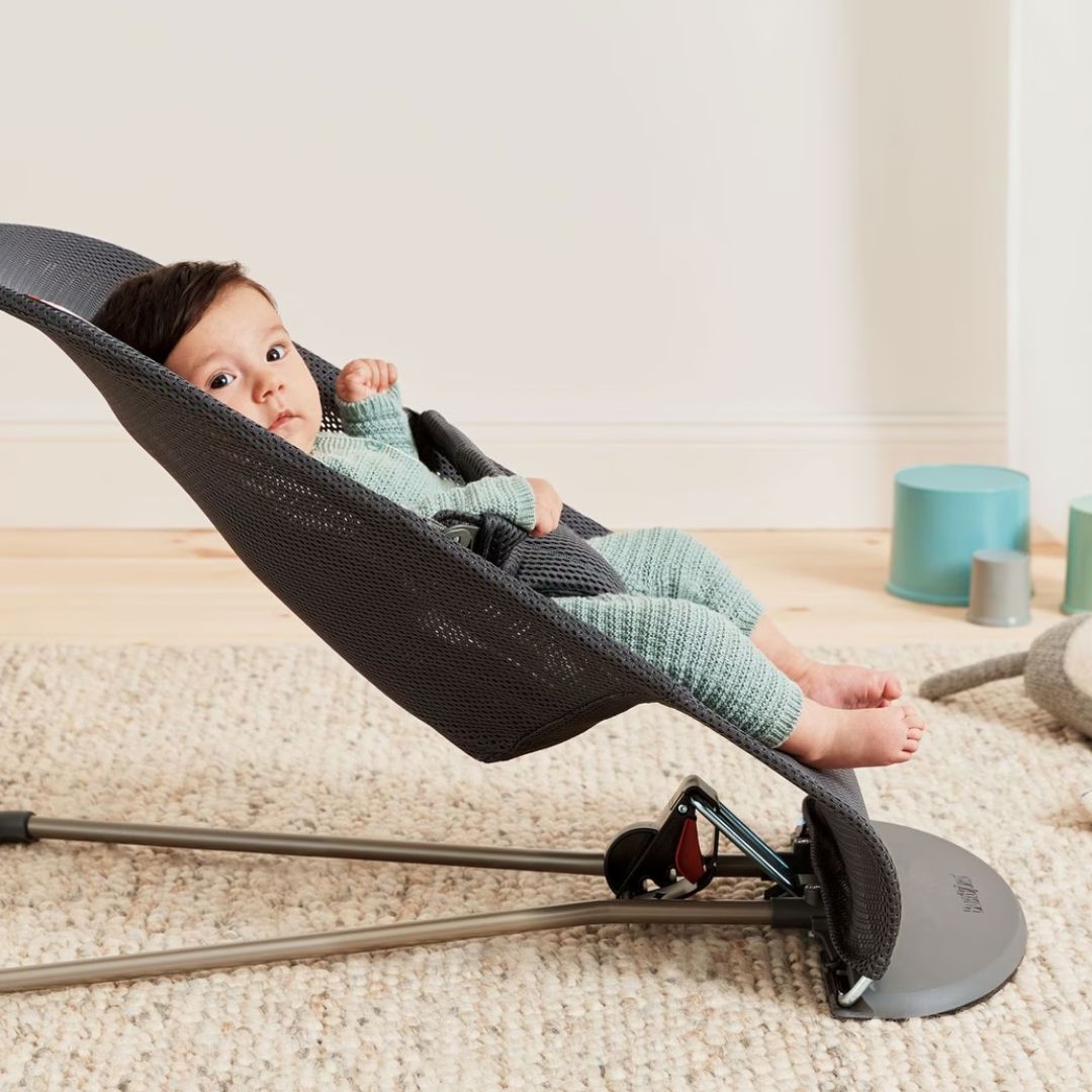 Baby in a baby jorn black baby bouncer on a carpeted floor with a neutral background
