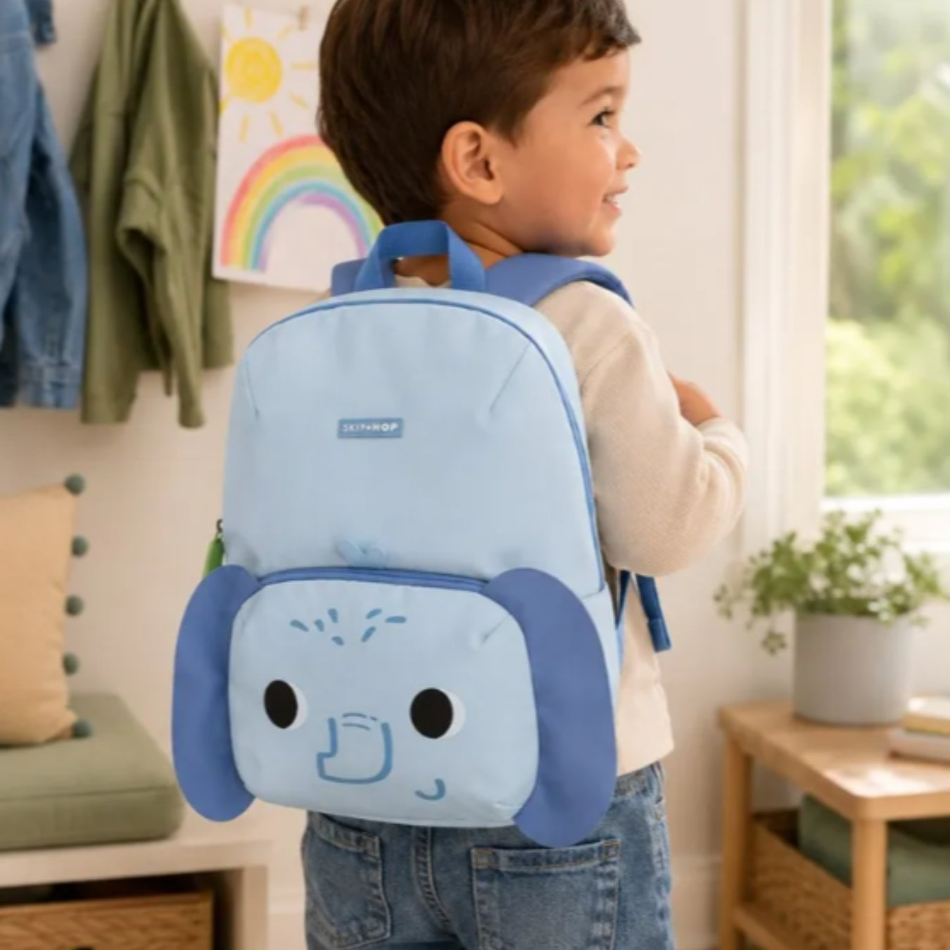 A young child wearing a light blue cartoon dog backpack in a bright, modern room. The Skip Hop backpack features a light blue body with darker blue floppy ears and a stylized Elephant face on the front pocket.