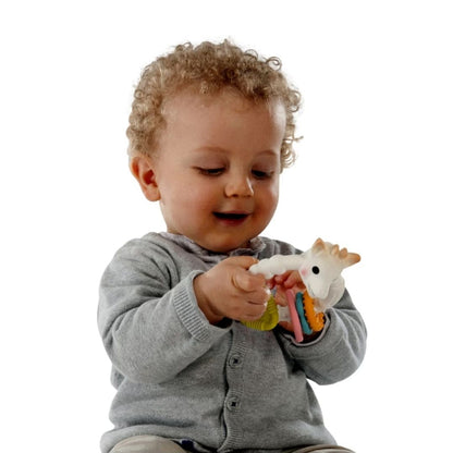 A curly-haired baby in a gray cardigan smiling while holding and playing with the Sophie la Girafe Colo'rings teething toy.