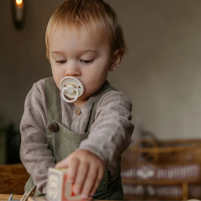 Child wearing a Frigg pacifier and reaching out with a box in a warm-toned room.