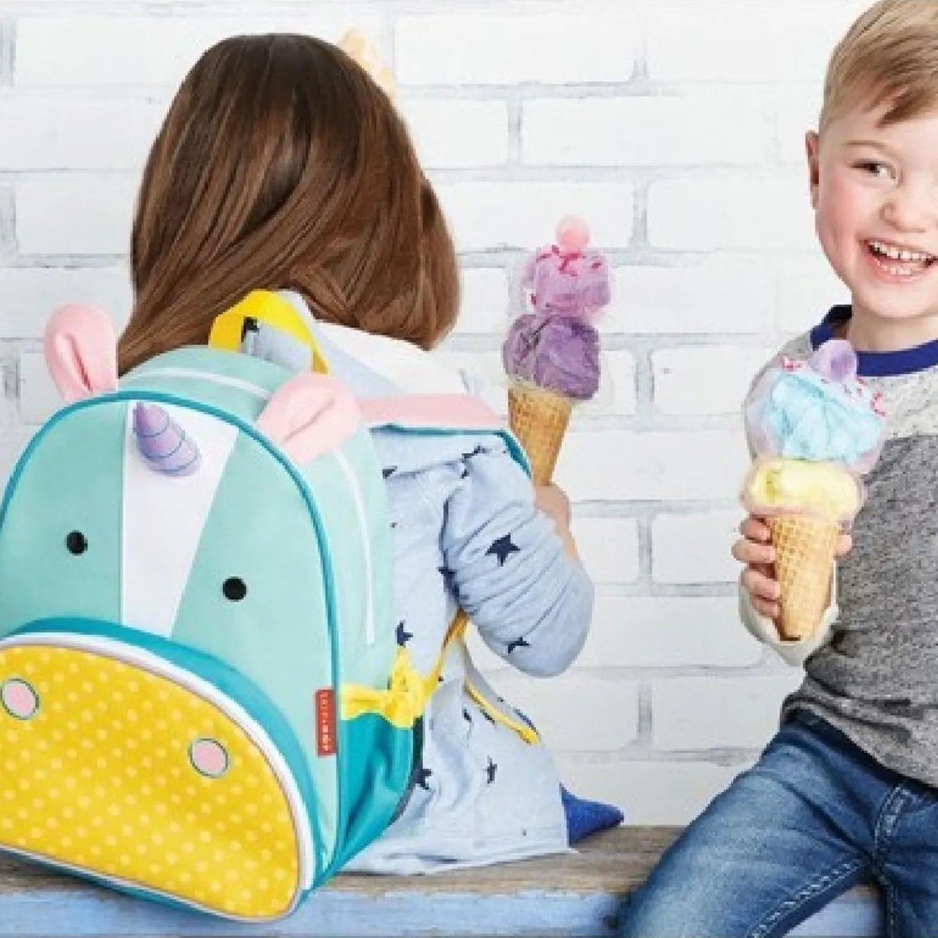 A child wearing a pastel unicorn Skip Hop backpack sits with a toy ice cream cone, while another child holds a colorful ice cream cone in front of a white brick wall.