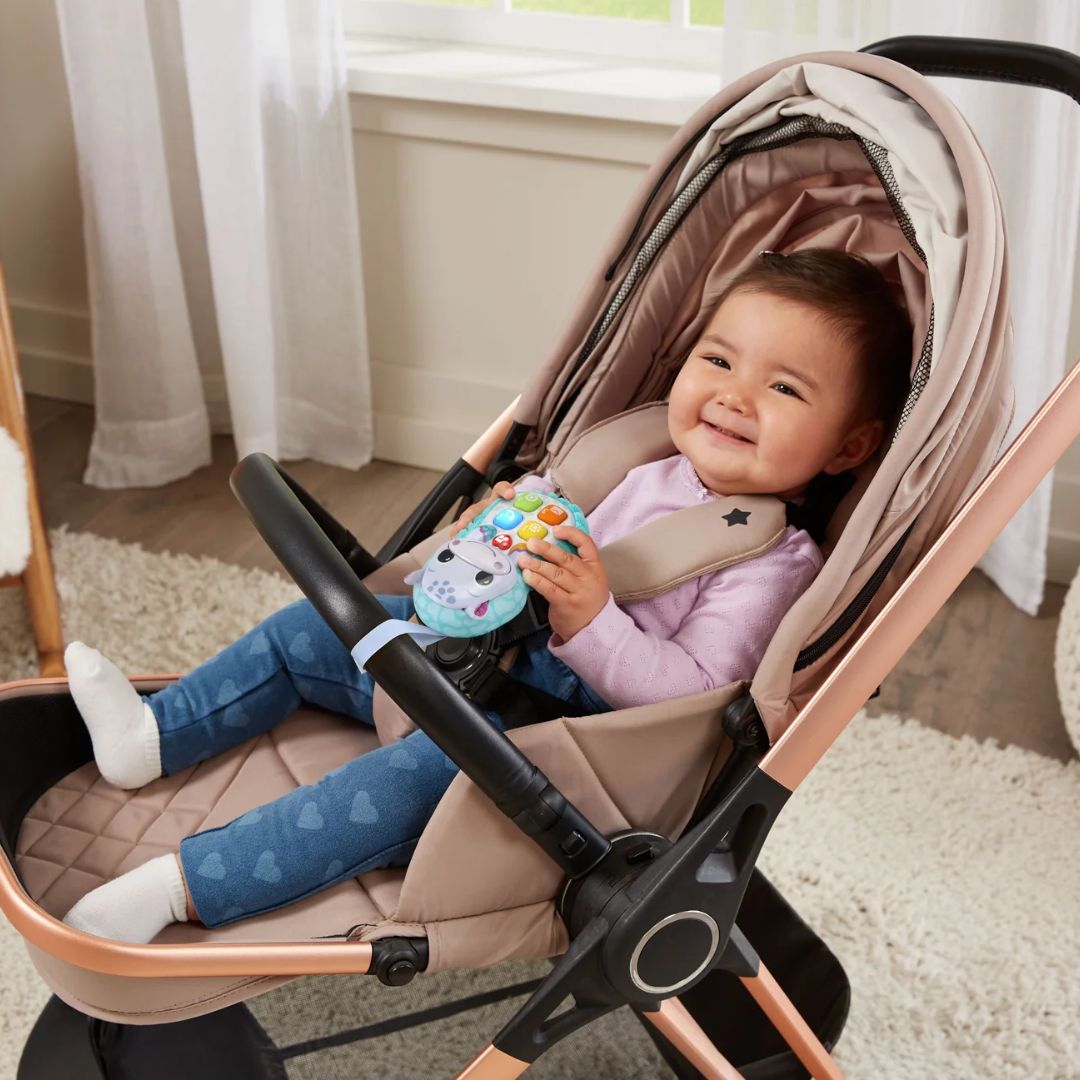 Child sitting in a stroller holding a Vtech toy, smiling indoors.