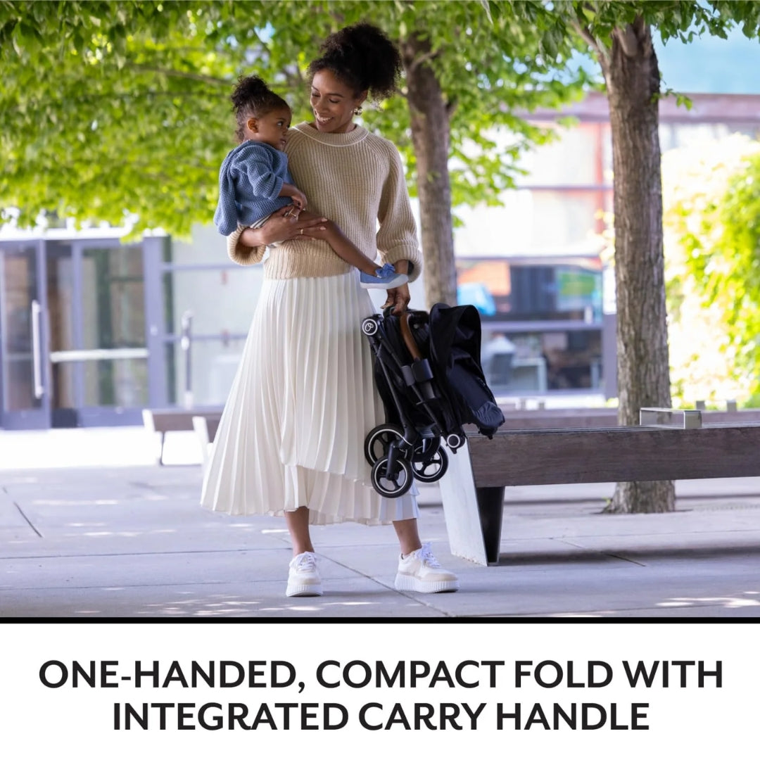 Woman holding a child and a compact Evenflo  stroller on a sidewalk with trees in the background.