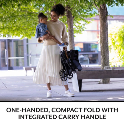 Woman holding a child and a compact Evenflo  stroller on a sidewalk with trees in the background.