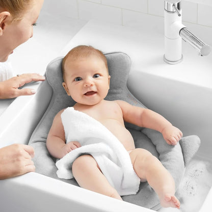 Baby sitting on a gray bath seat with a white towel, being bathed by an adult in a sink.