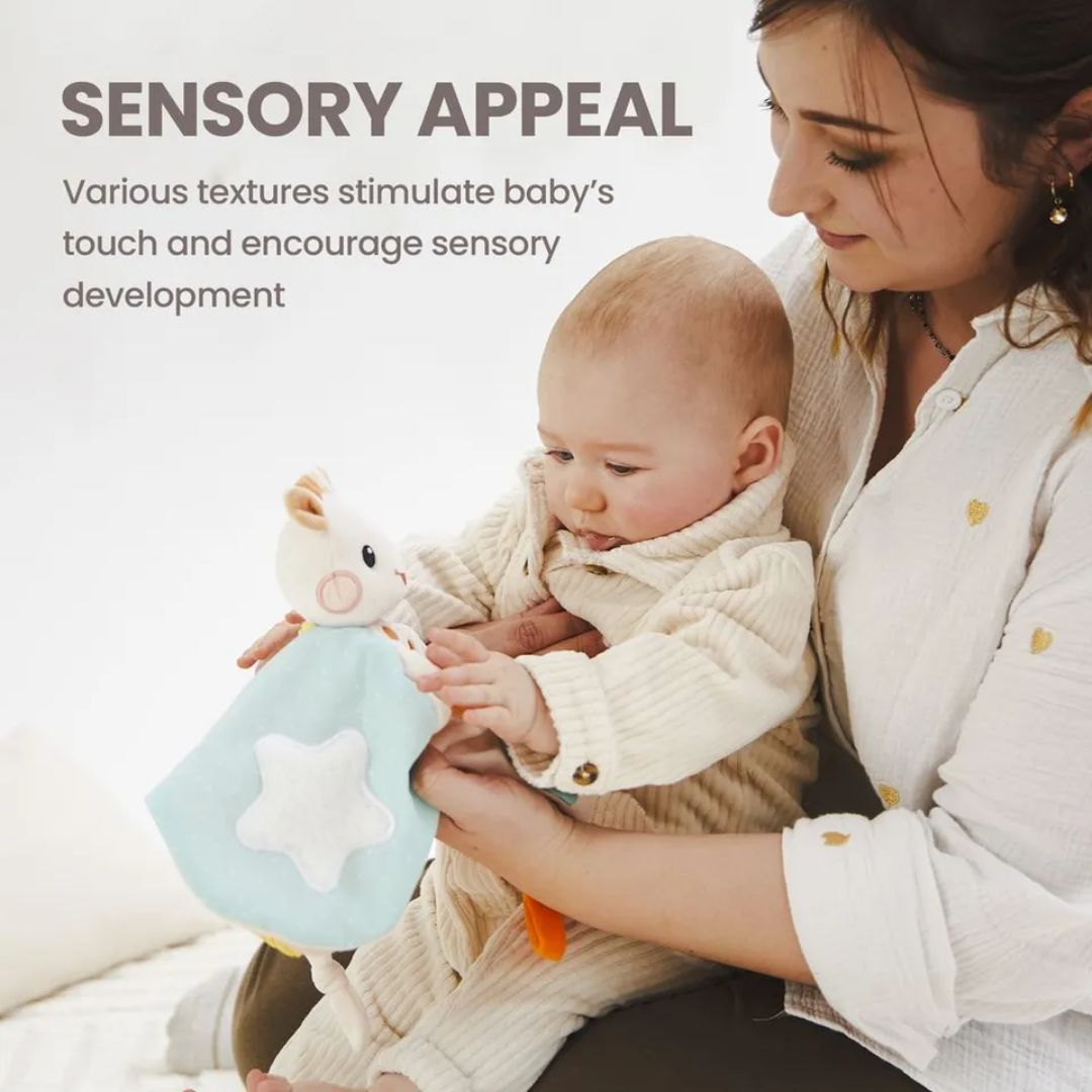 A mother holding her baby who is touching a blue star-patterned Sophie la Girafe comforter, with text reading 'Sensory Appeal: Various textures stimulate baby's touch and encourage sensory development.