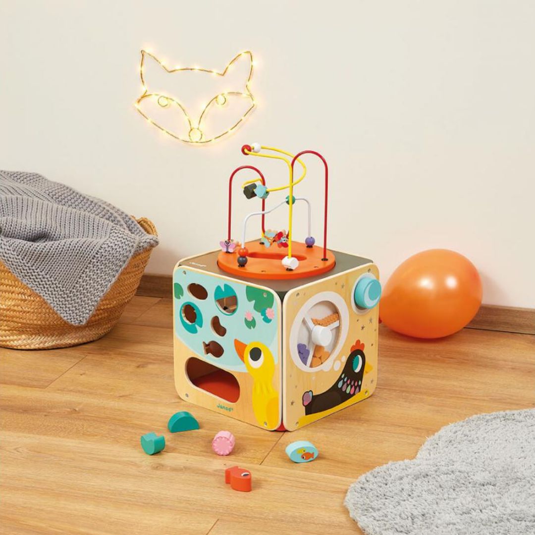 Colorful Janod wooden toy cube with beads and shapes on a wooden floor.