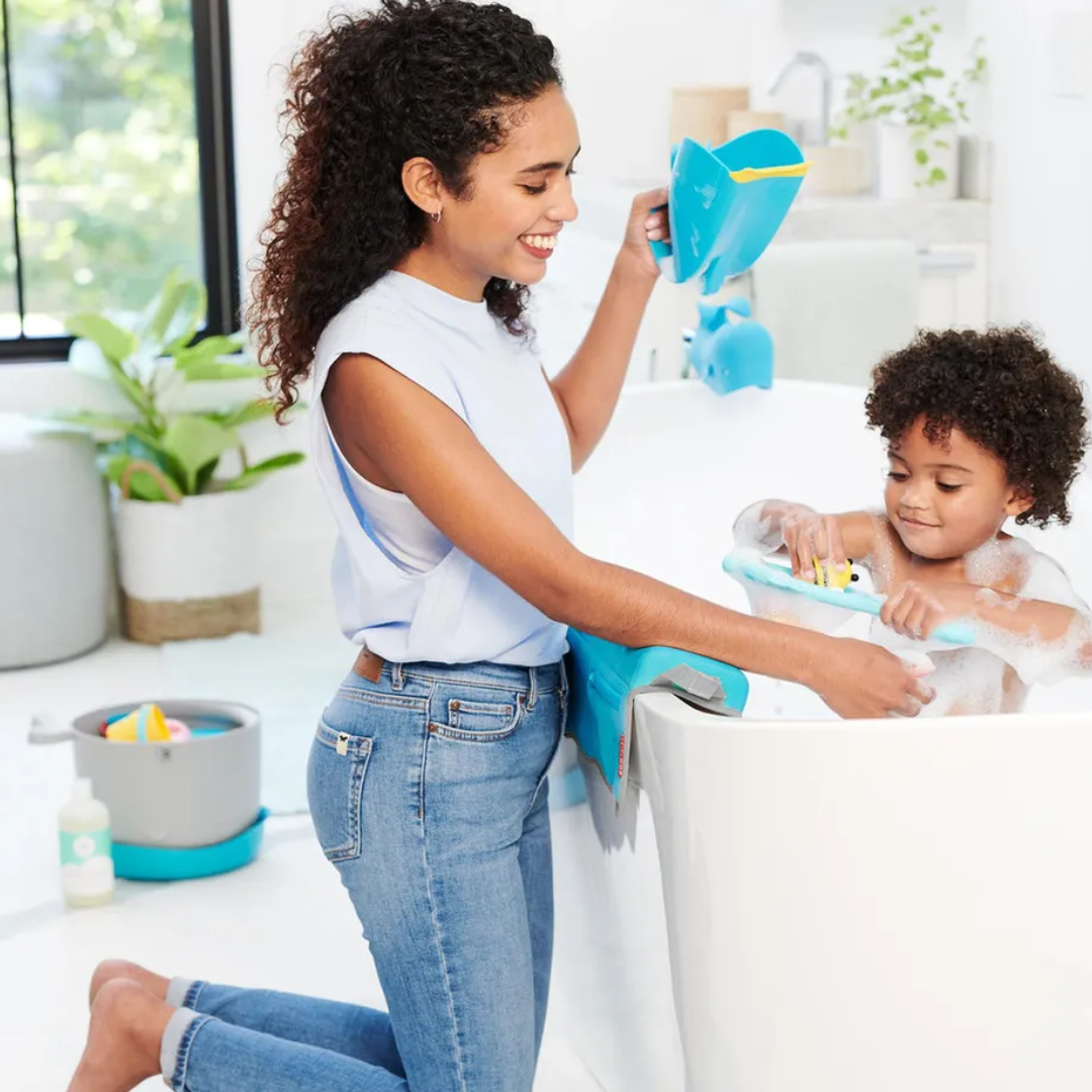 A smiling mother kneeling beside the bathtub holding the skip hop blue whale-shaped bath rinser, while her toddler plays with bath toys in the bubbly water, with a small storage caddy and bath products nearby.