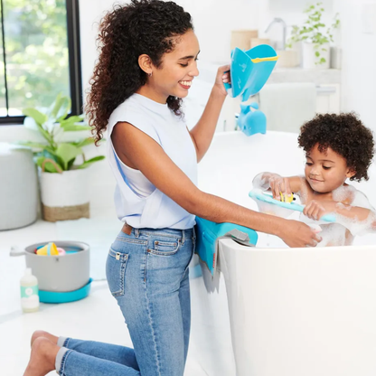 A smiling mother kneeling beside the bathtub holding the skip hop blue whale-shaped bath rinser, while her toddler plays with bath toys in the bubbly water, with a small storage caddy and bath products nearby.