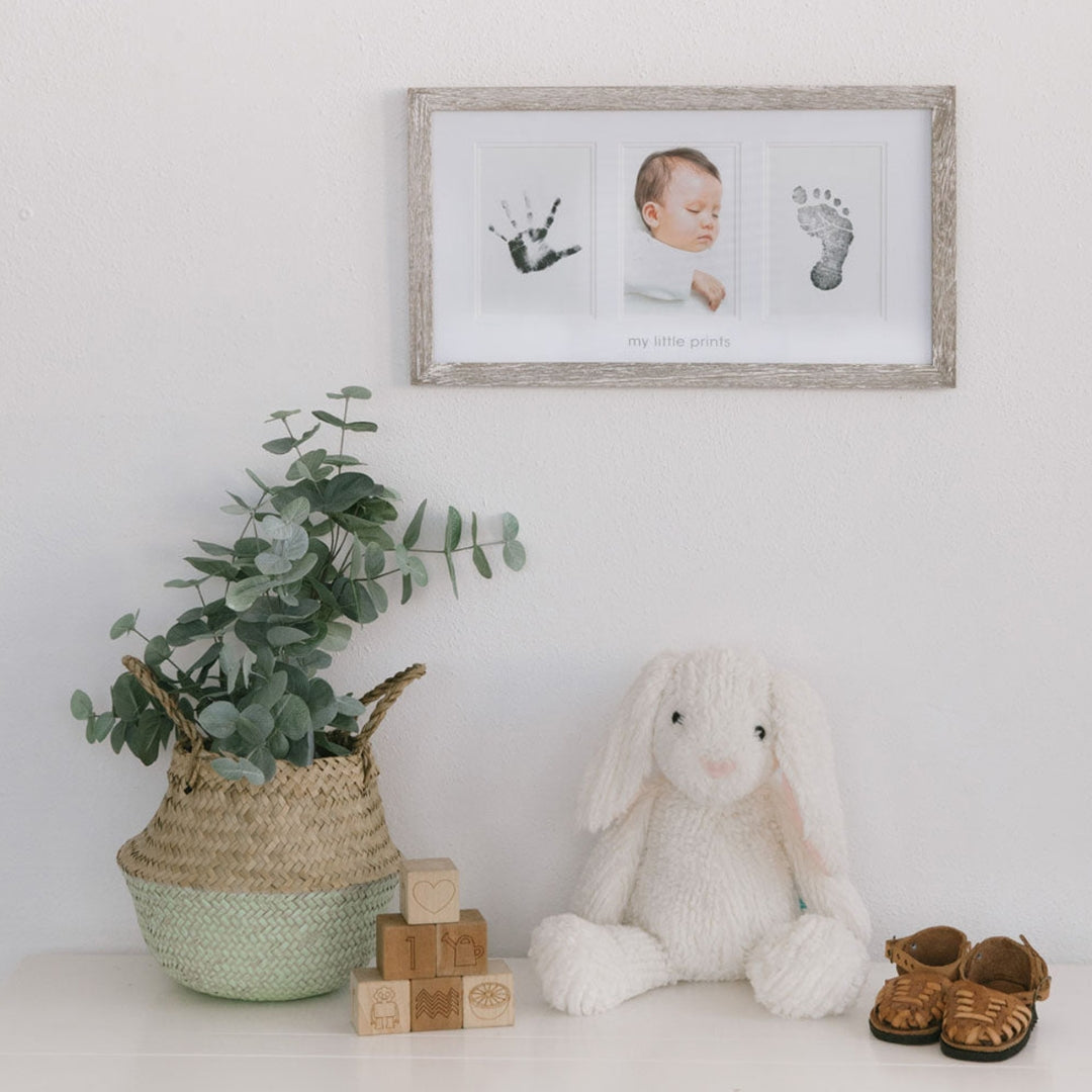 Framed baby milestone photos with a stuffed bunny, plant, and shoes on a white surface.