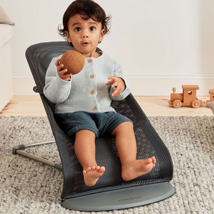 Child sitting on a Baby Jorn baby bouncer holding a ball in a room with toys.