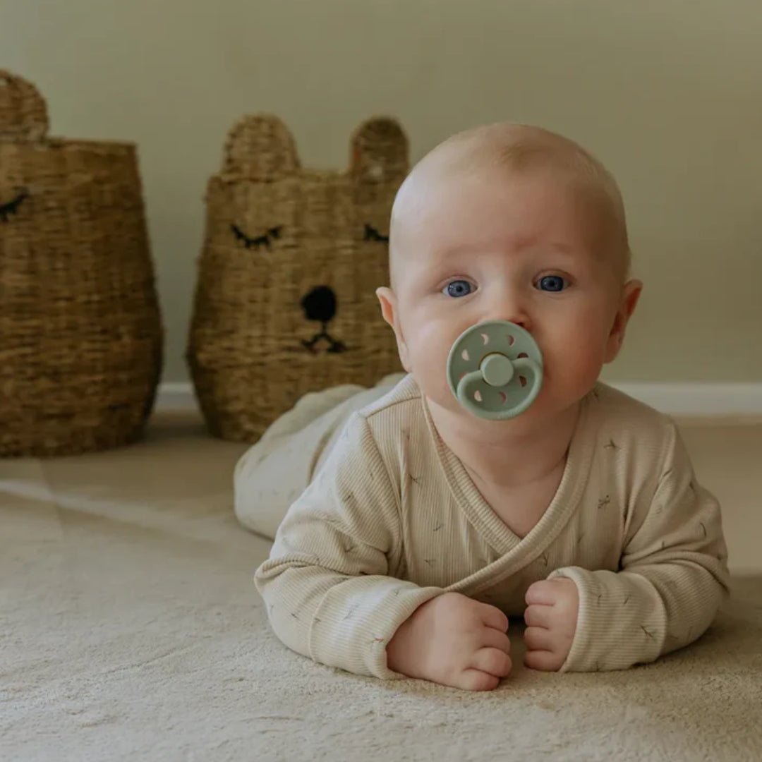 Baby lying on a blanket with a pacifier, surrounded by wicker baskets in the background.