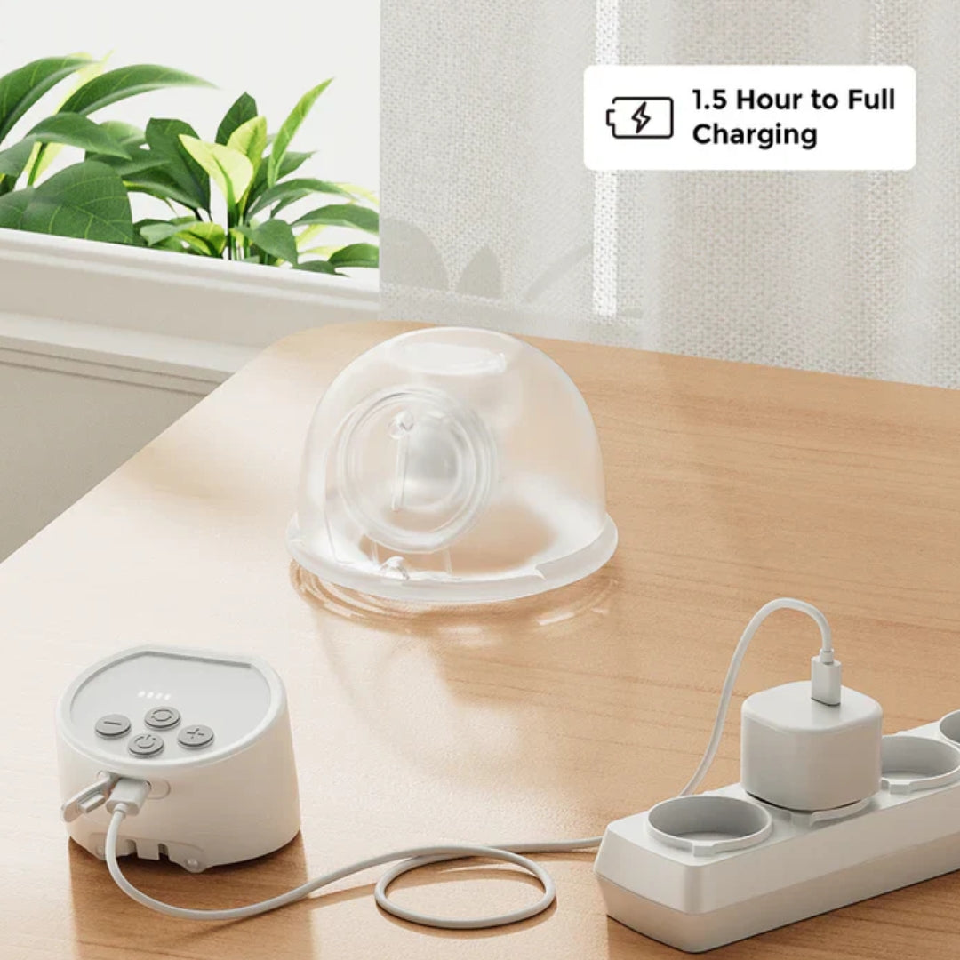White Momcozy electronic wearable breast pump with charging cable on a wooden surface, plant and window in the background.