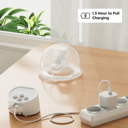White Momcozy electronic wearable breast pump with charging cable on a wooden surface, plant and window in the background.