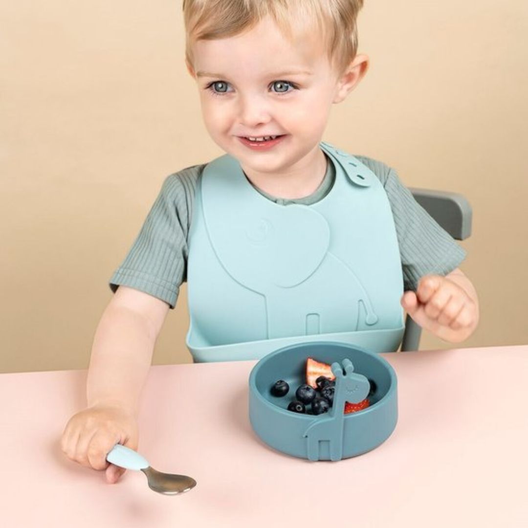 Child wearing a light blue bib and eating from a bowl with a spoon on a pink table.